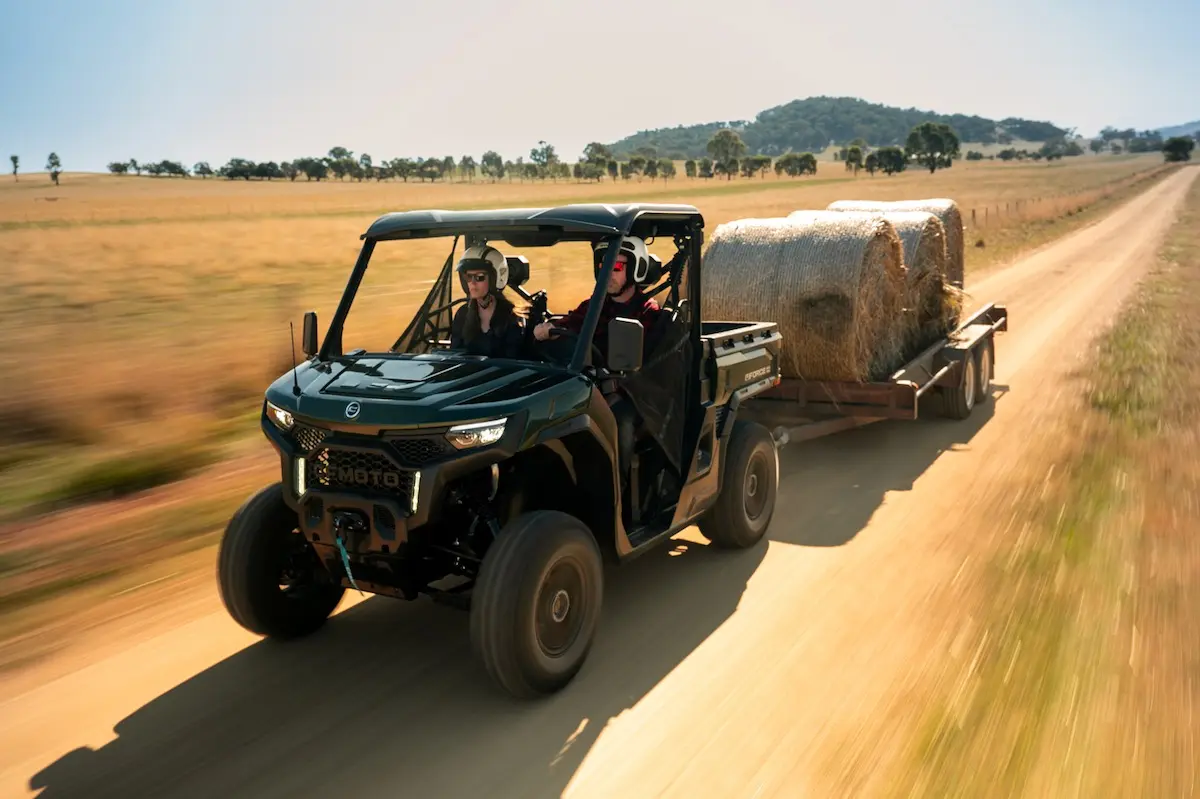 A UFORCE U10 PRO AGRI utility vehicle towing a trailer loaded with large hay bales drives down a rural dirt road on a sunny day.