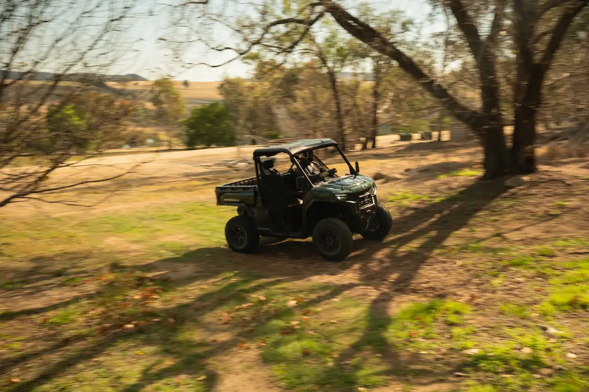 A green UFORCE U10 PRO AGRI utility terrain vehicle drives across a grassy, lightly wooded area with trees casting shadows and open fields visible in the background.