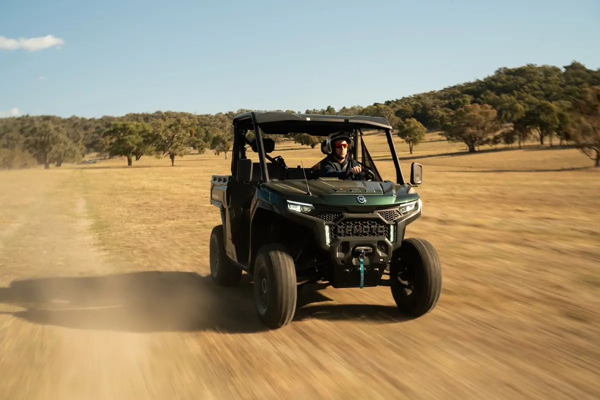 A person drives a dark green UFORCE U10 PRO AGRI utility terrain vehicle across a dry, grassy field with trees and hills in the background.