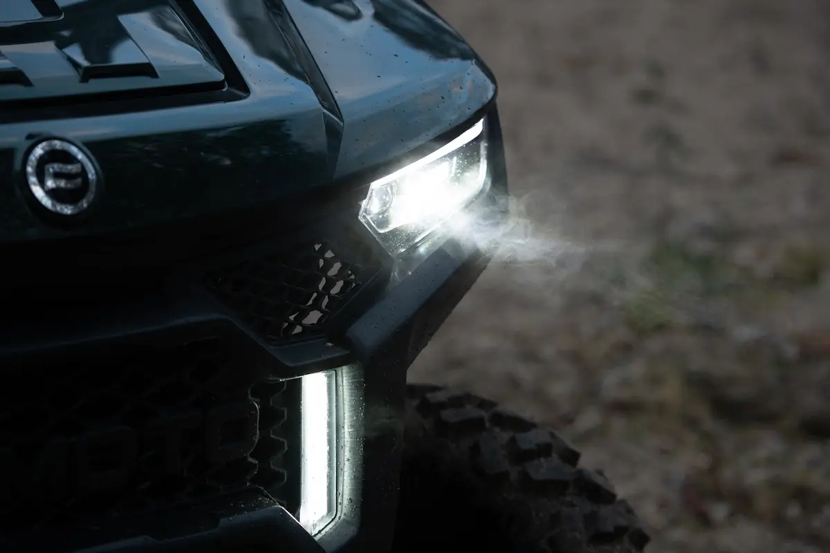 Close-up of the front left headlight and grille of the UFORCE U10 PRO AGRI off-road vehicle with headlights turned on, emitting light in a low-light outdoor setting.