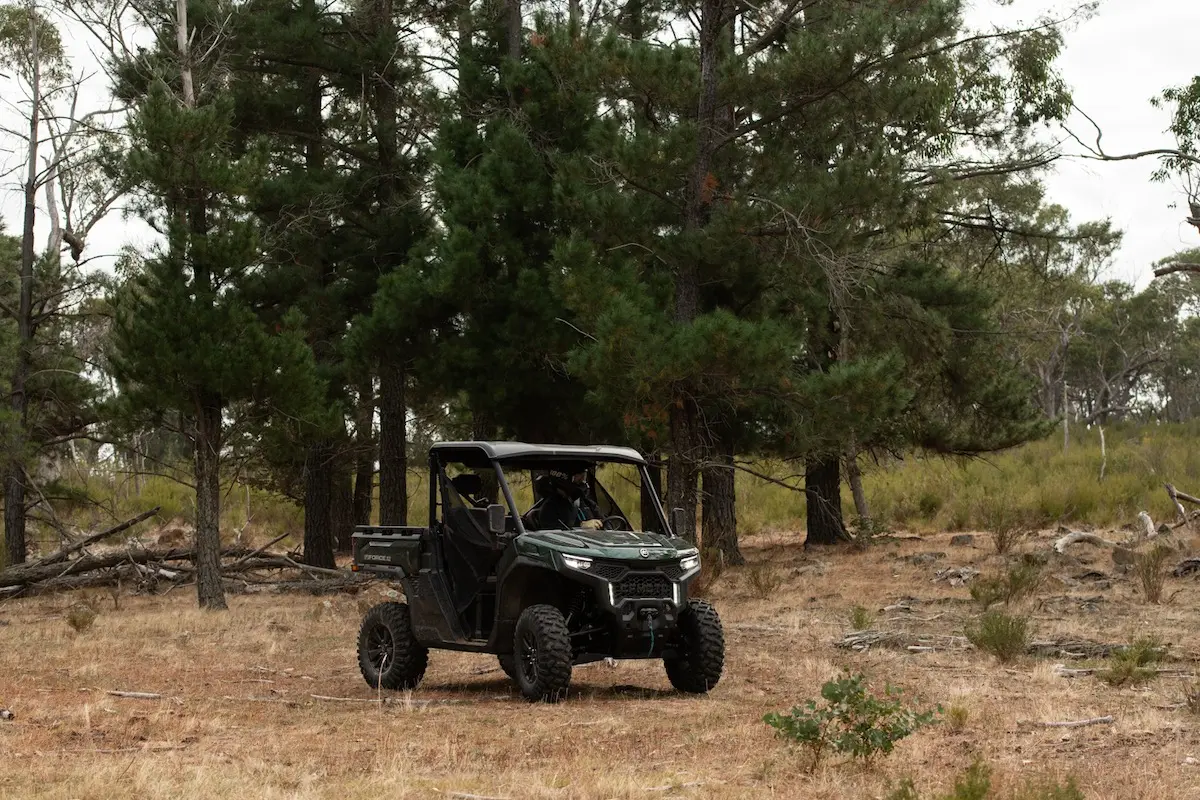 A black UFORCE U10 PRO AGRI all-terrain utility vehicle is parked on dry grass in front of tall pine trees in a forested area.