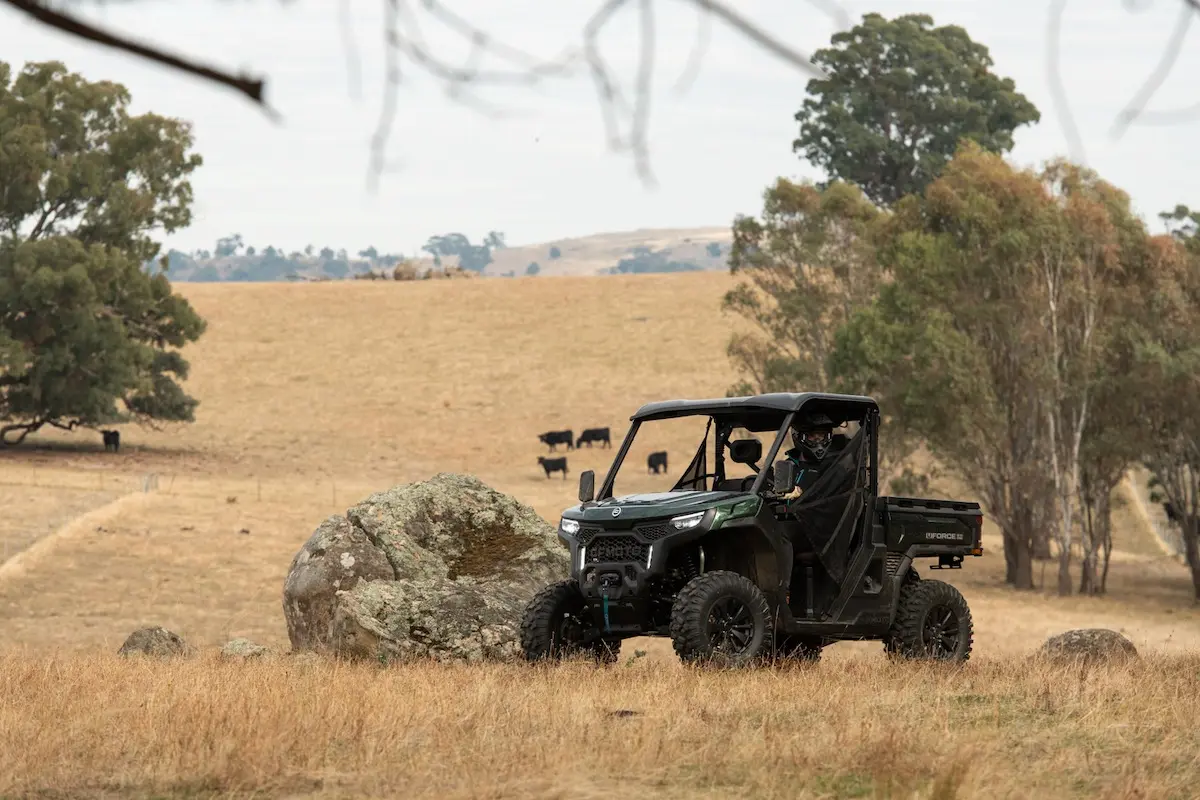 A black UFORCE U10 PRO AGRI side-by-side utility vehicle with two occupants drives across a dry, grassy field with scattered trees, rocks, and grazing cattle in the background.