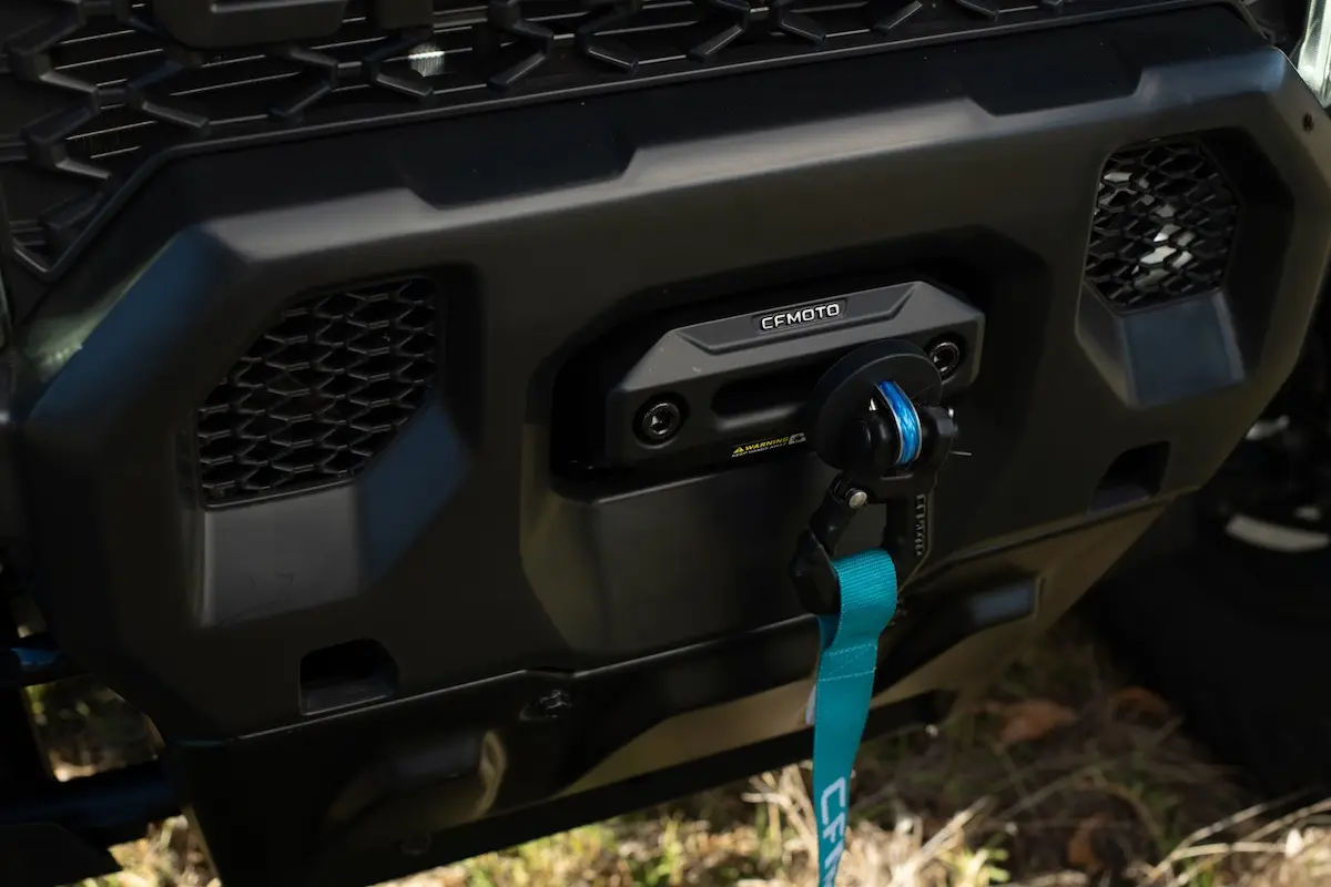 Close-up of a CFMOTO UFORCE U10 PRO AGRI vehicle winch mounted on the front bumper with a blue tow strap attached, surrounded by a black grille and bodywork.