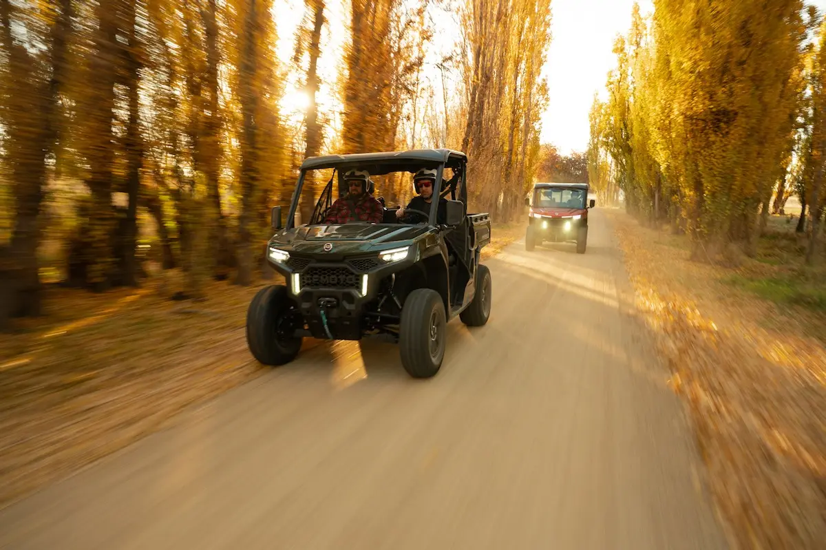 Two UFORCE U10 PRO AGRI off-road utility vehicles drive down a dirt road surrounded by tall autumn trees, with sunlight streaming through the branches.