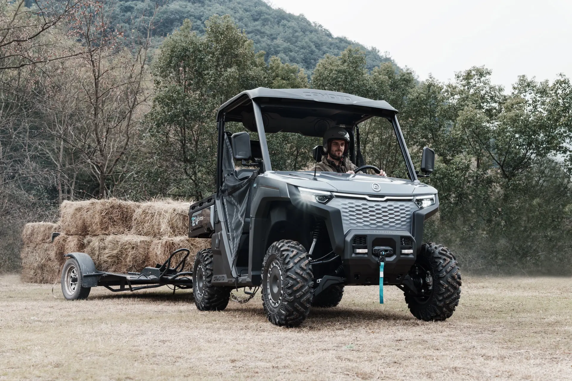 A man drives a UFORCE 600 utility terrain vehicle towing a trailer loaded with hay bales across a grassy field, with trees and hills in the background.