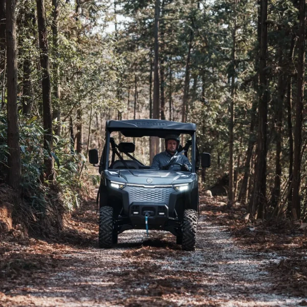 A person drives a UFORCE EVU6 Commercial off-road utility vehicle along a dirt path surrounded by tall trees in a forested area.