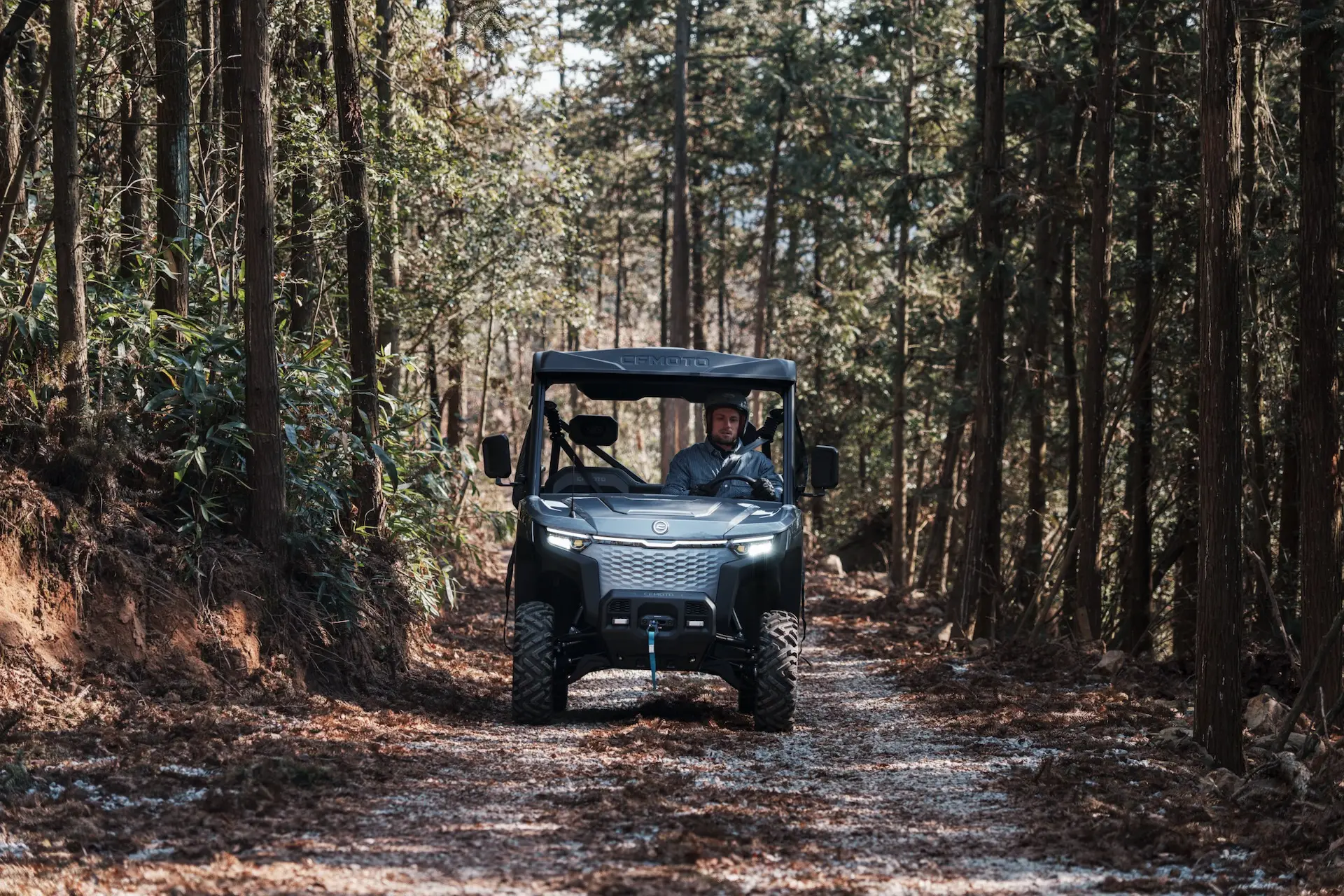 A man drives a black UFORCE 600 utility vehicle on a narrow dirt trail surrounded by tall trees in a forest.