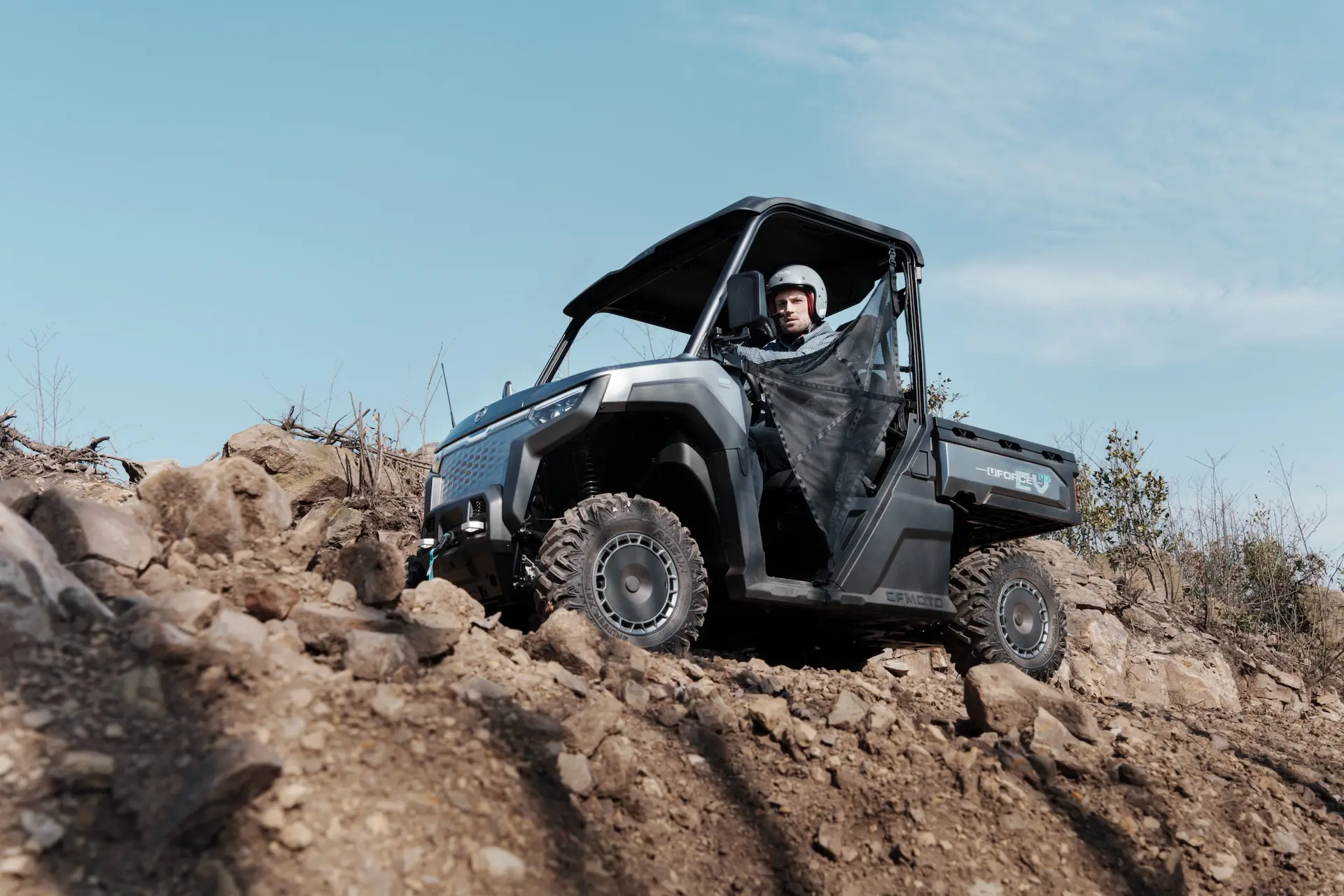 A person wearing a helmet drives a UFORCE 600 utility terrain vehicle (UTV) over rocky, uneven ground under a clear blue sky.