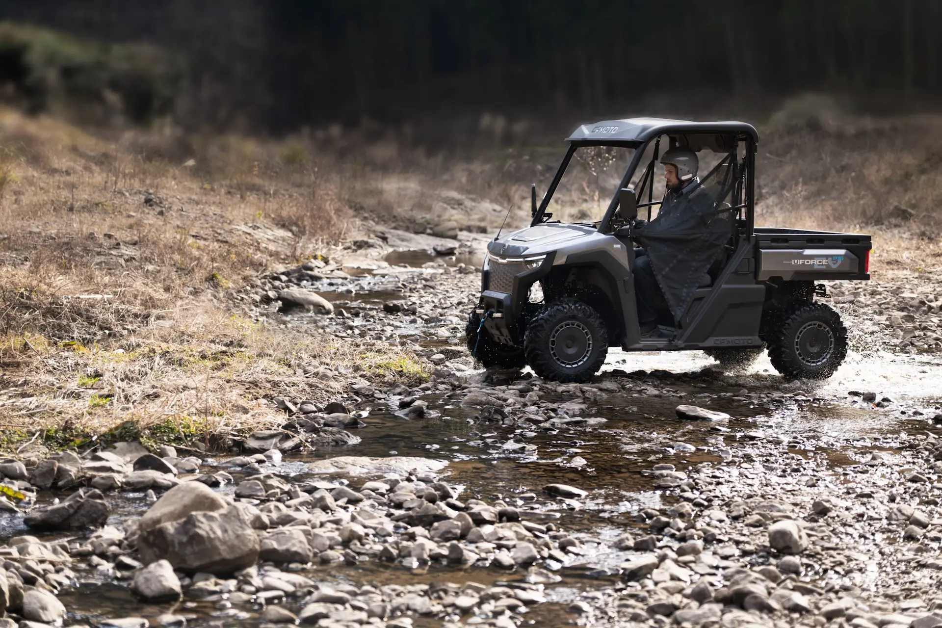 A person drives a UForce 600 off-road utility vehicle across a shallow, rocky stream in a dry, grassy landscape.
