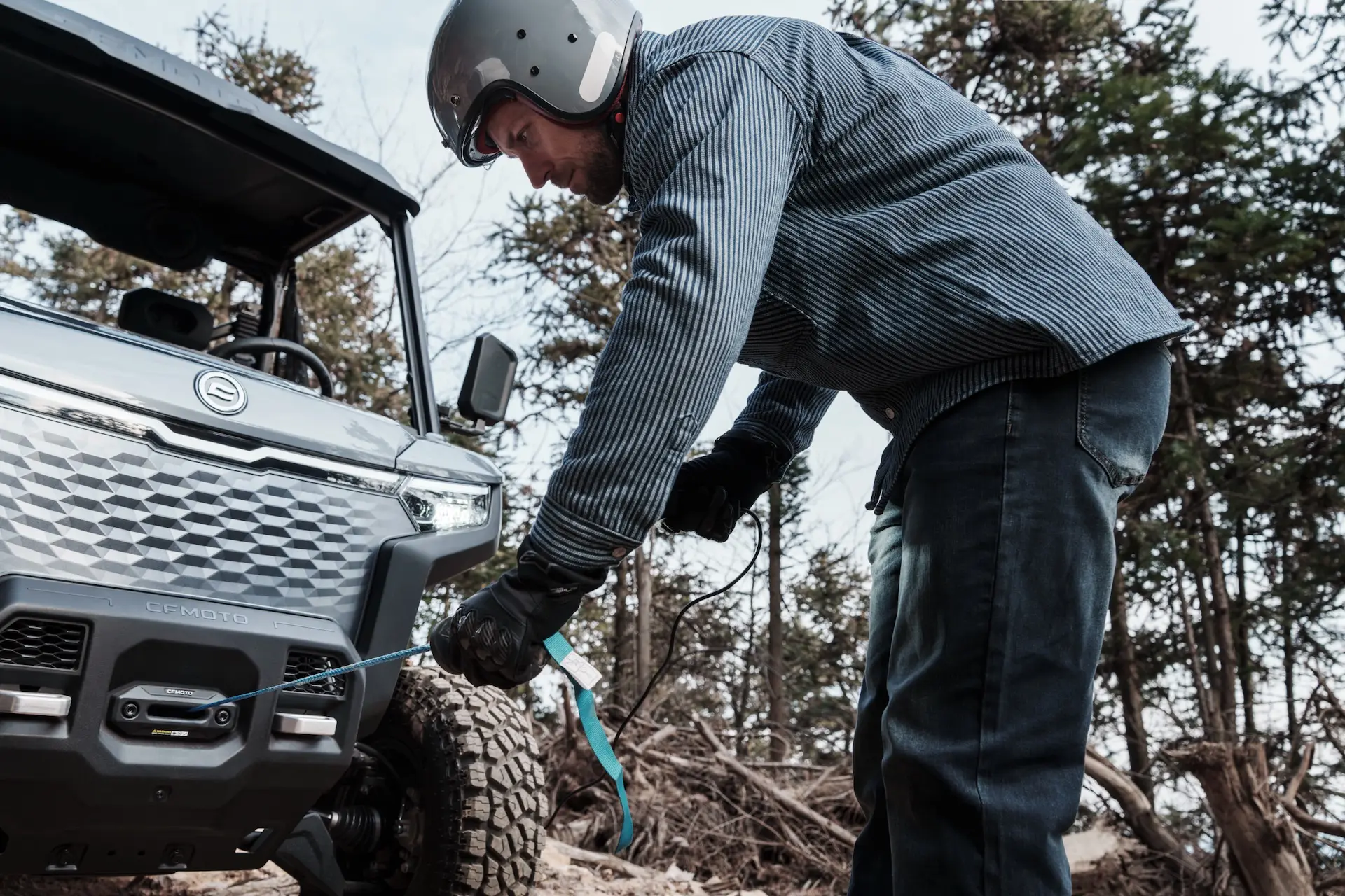 A person wearing a helmet and gloves attaches a tow strap to the front of a UFORCE 600 off-road vehicle in a wooded area.