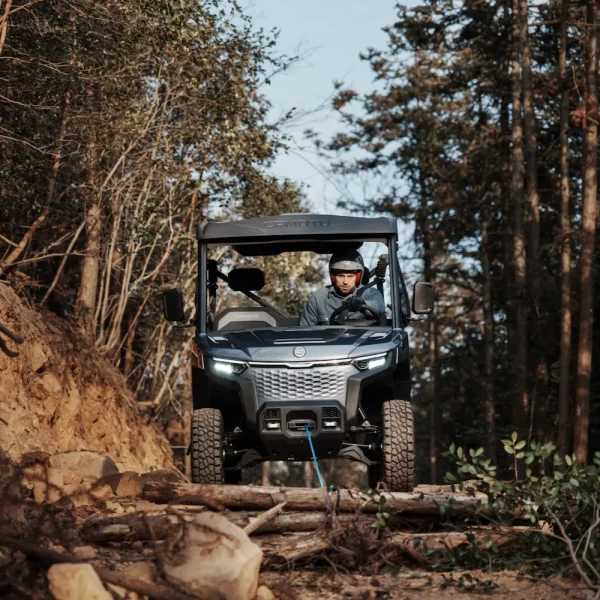 A person drives a UFORCE EVU6 Commercial over a fallen tree on a dirt trail surrounded by trees.