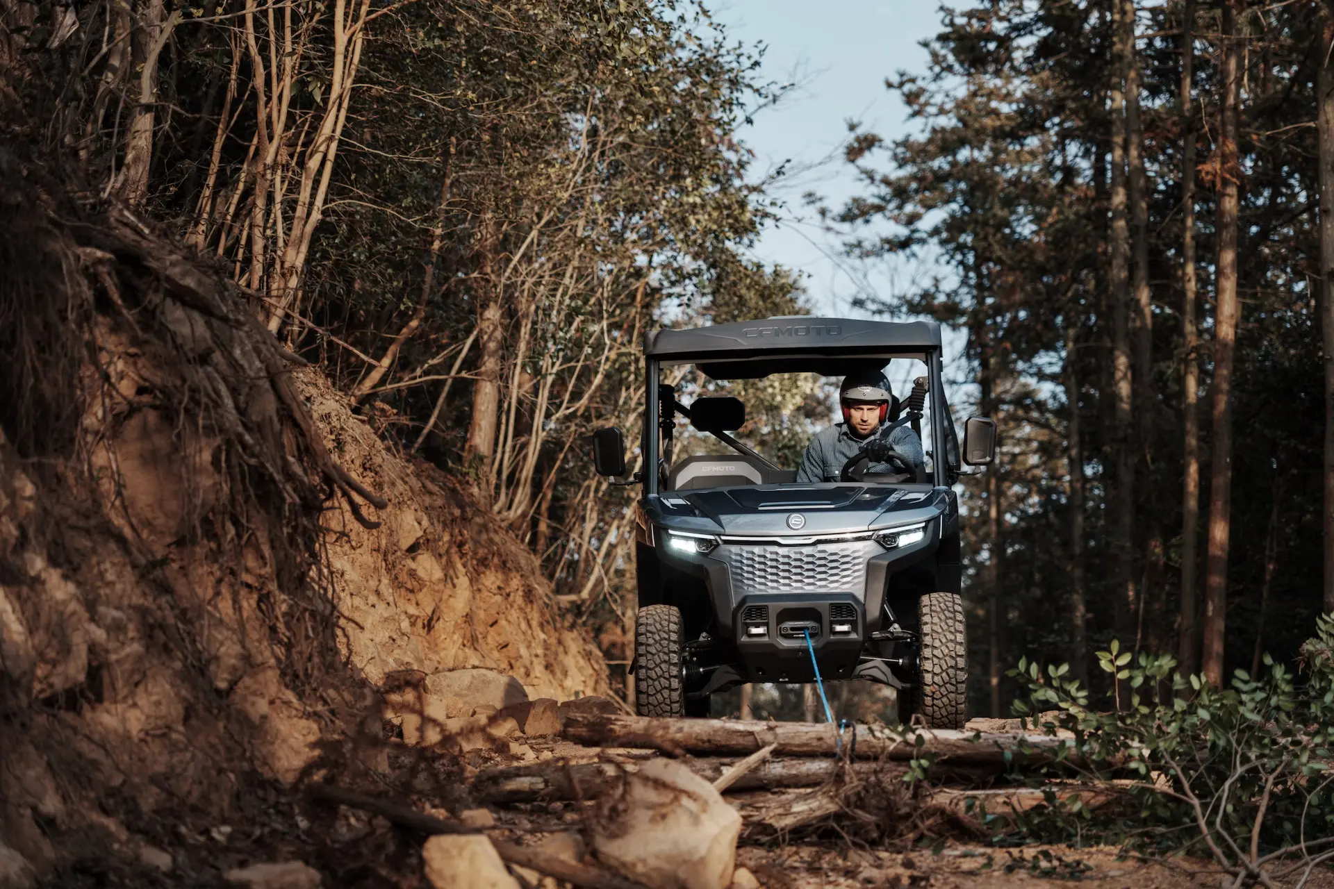 A person drives a UFORCE 600 off-road vehicle down a rocky, wooded trail with fallen branches and uneven terrain.