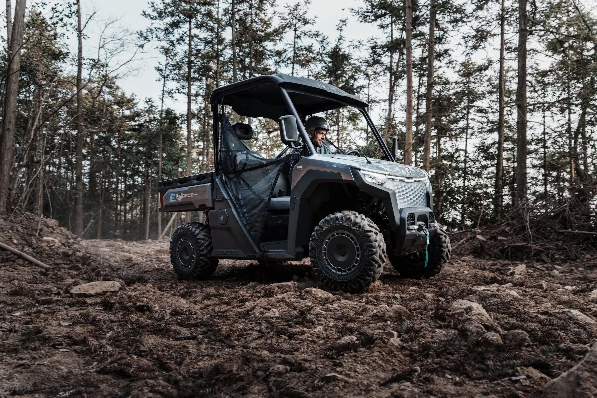 A man drives a black UFORCE 600 utility terrain vehicle on a rocky, dirt path surrounded by trees in a forested area.