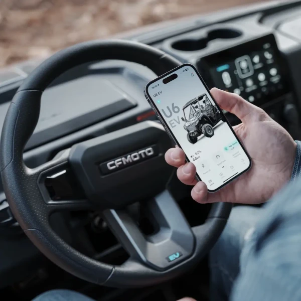 A person holds a smartphone displaying a vehicle app inside a CFMOTO UFORCE EVU6 Commercial, with the steering wheel and dashboard visible.