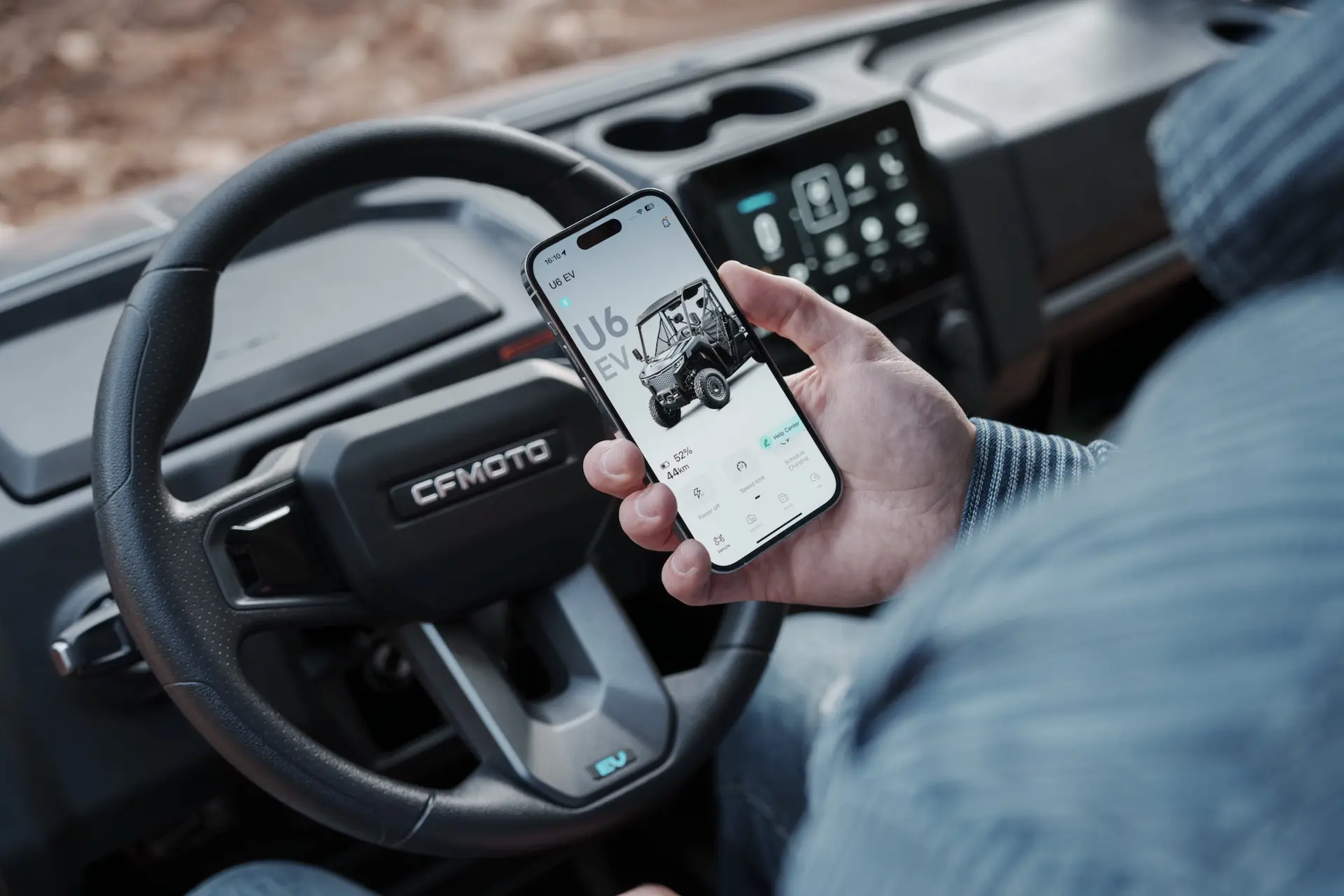 A person holding a smartphone with a vehicle app open, sitting in the driver’s seat of a CFMOTO UFORCE 600 with the dashboard visible.