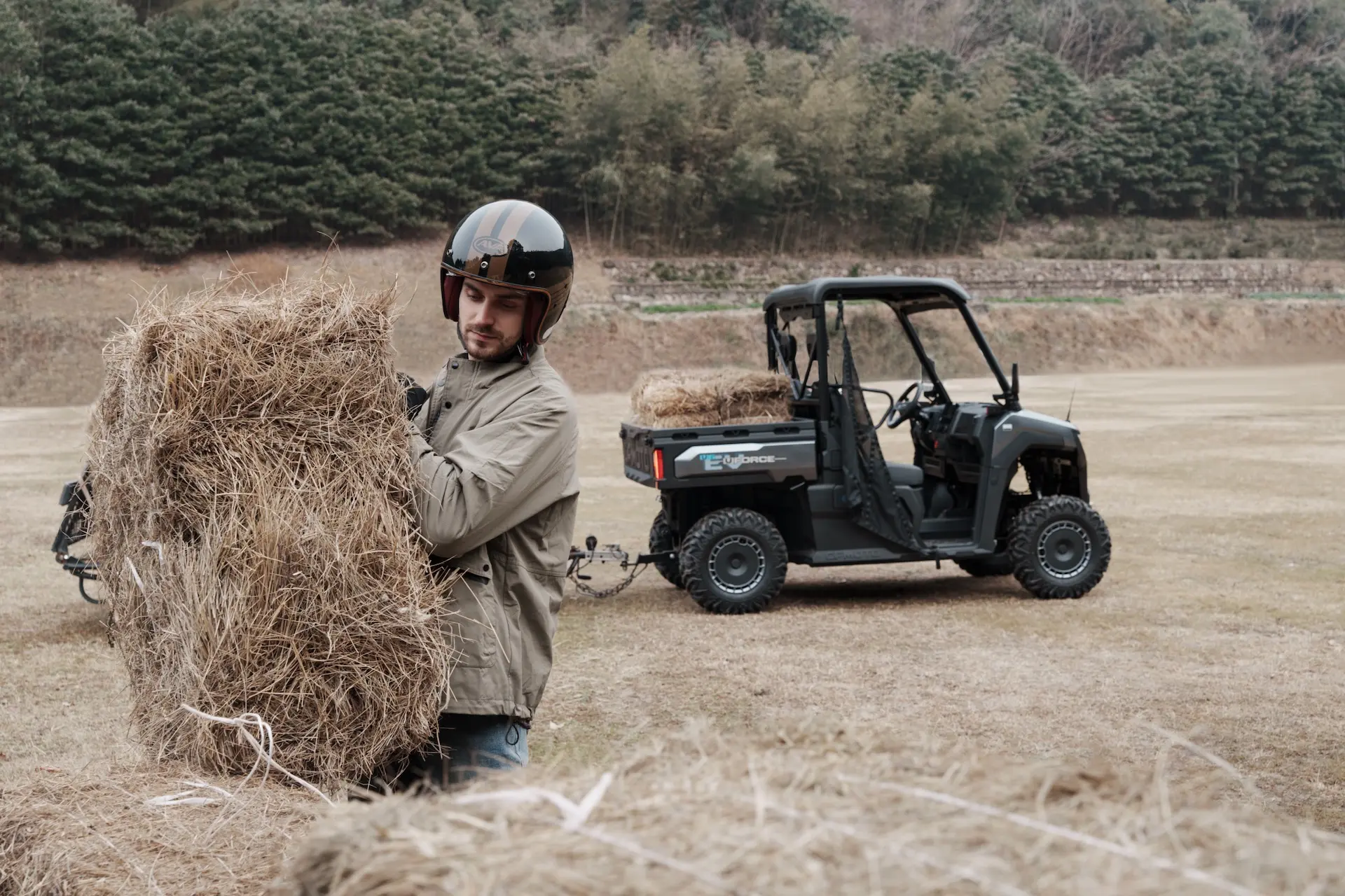 A person wearing a helmet and jacket lifts a bale of hay in a field, with a UFORCE 600 off-road utility vehicle carrying more hay in the background.