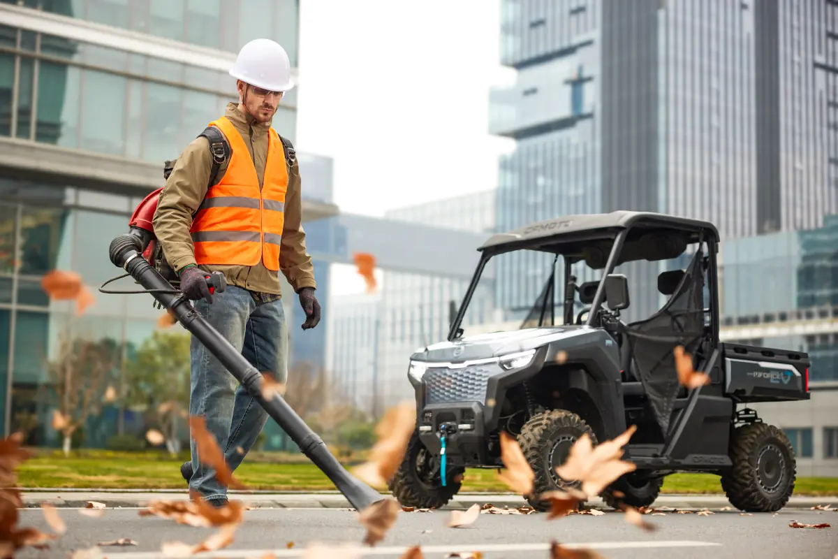 A worker in a helmet and safety vest uses a leaf blower on a city pavement near a parked electric UTV, with buildings in the background.