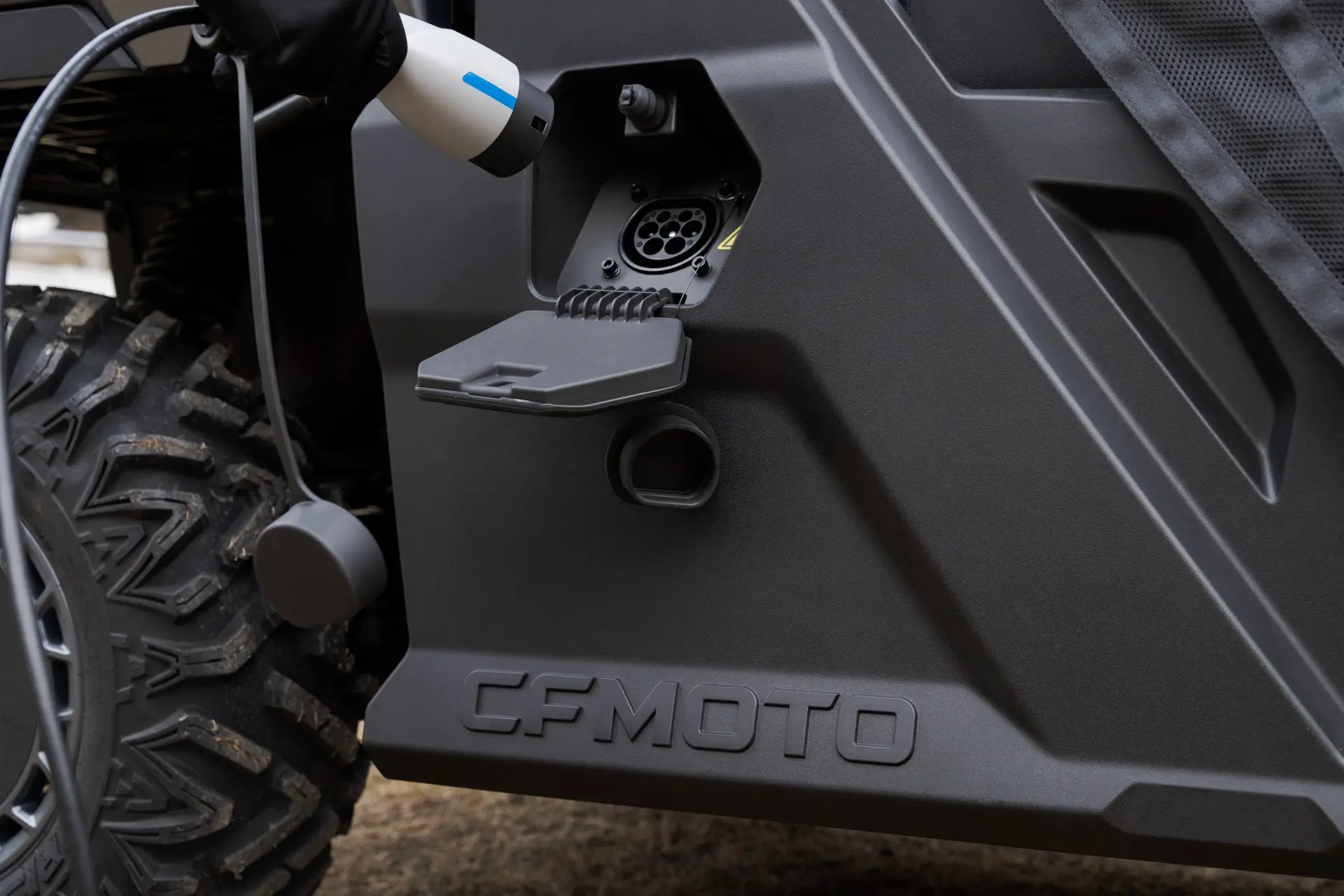 A charging cable is plugged into the charging port of a CFMOTO UFORCE 600 electric vehicle, shown near the rear wheel and side panel.