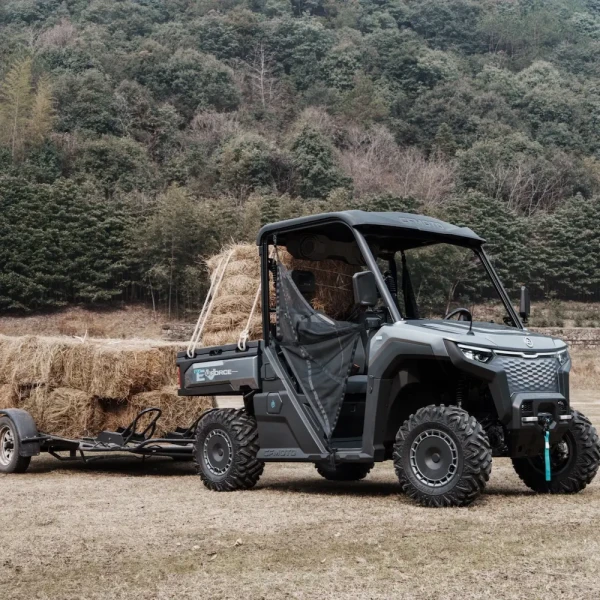 A UFORCE EVU6 Commercial utility vehicle with large tires is towing a trailer loaded with hay bales in a grassy area, set against a forested hillside backdrop.