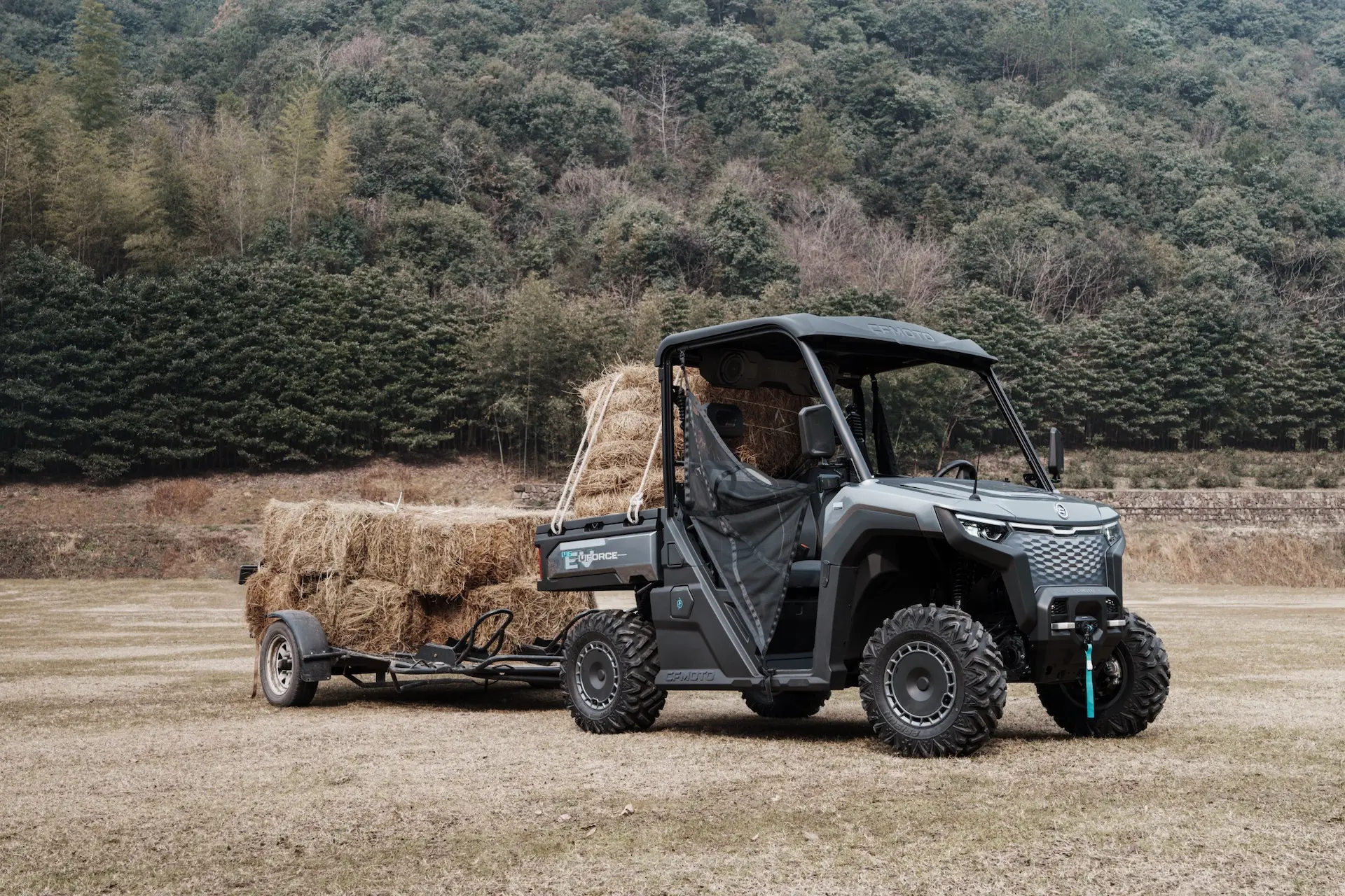 A uforce 600 utility vehicle pulls a trailer loaded with hay bales across a grassy field, with dense green trees in the background.