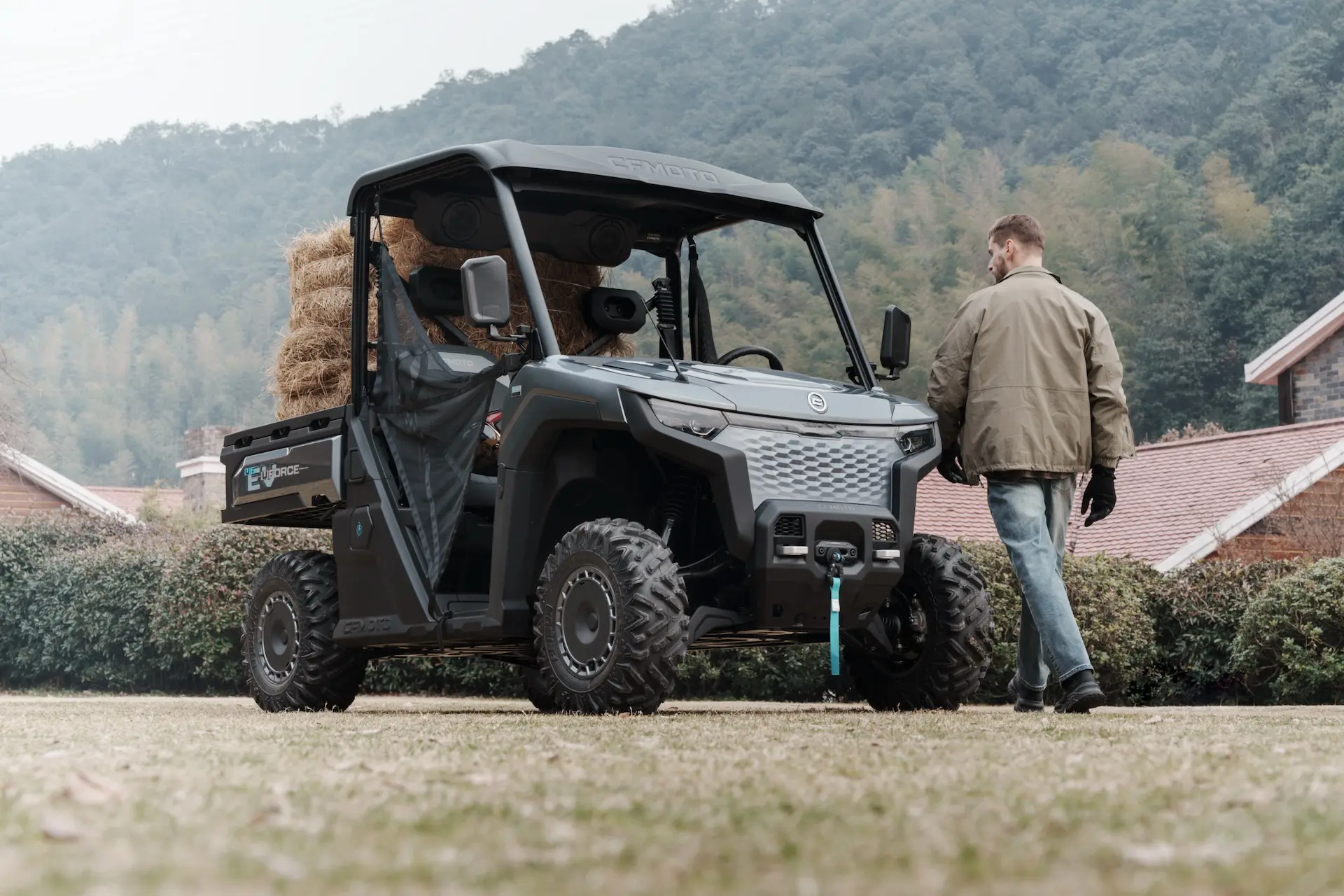 A man stands next to a black UFORCE 600 utility vehicle loaded with hay bales, parked on grass with hills and buildings in the background.