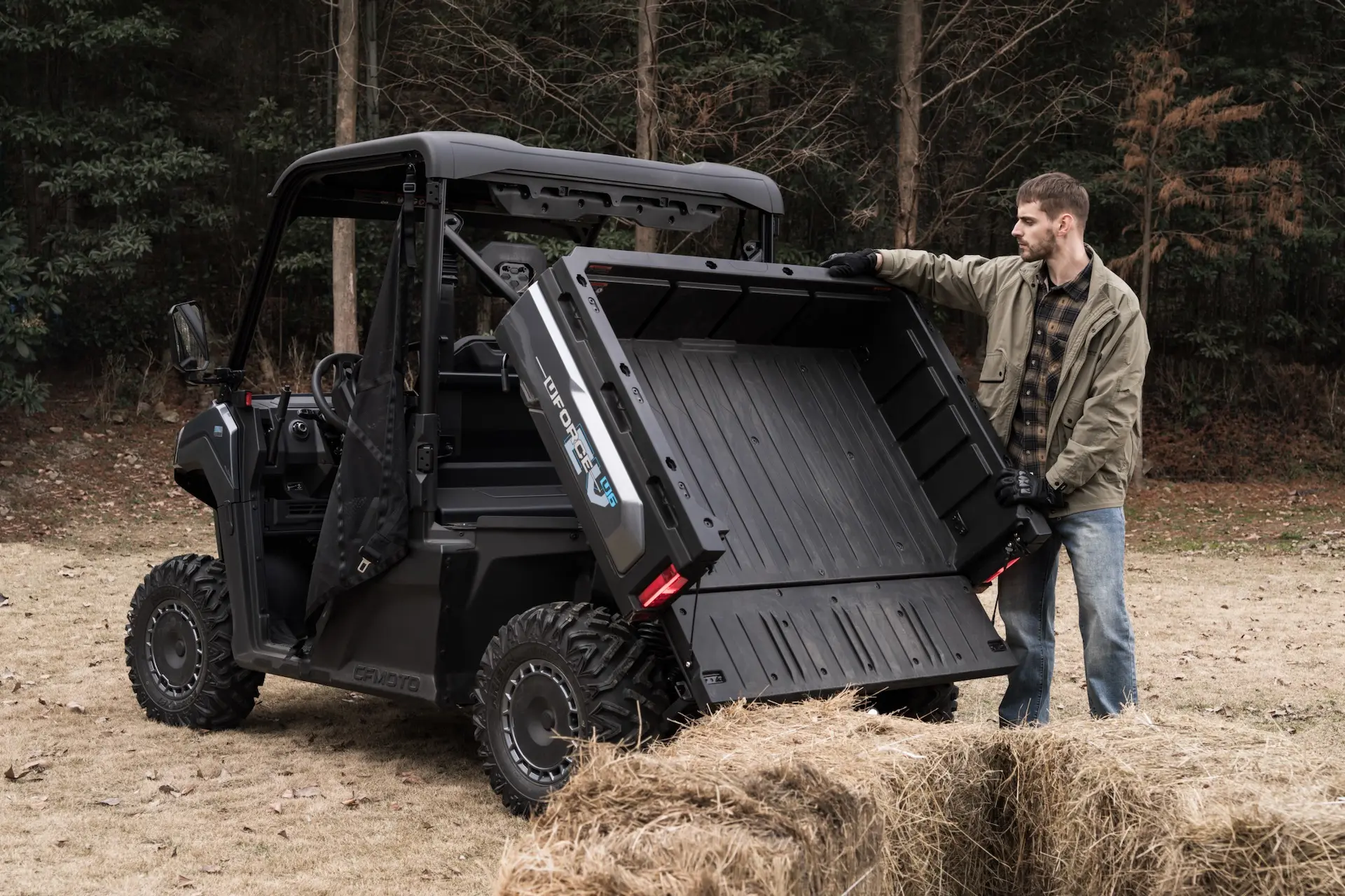 A person stands next to a black UFORCE 600 utility vehicle with its cargo bed tilted, unloading hay bales in an outdoor setting.