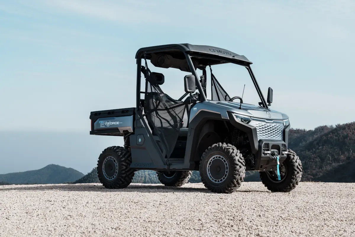 A modern electric UTV with off-road tyres is parked on gravel with mountains in the background under a clear sky.