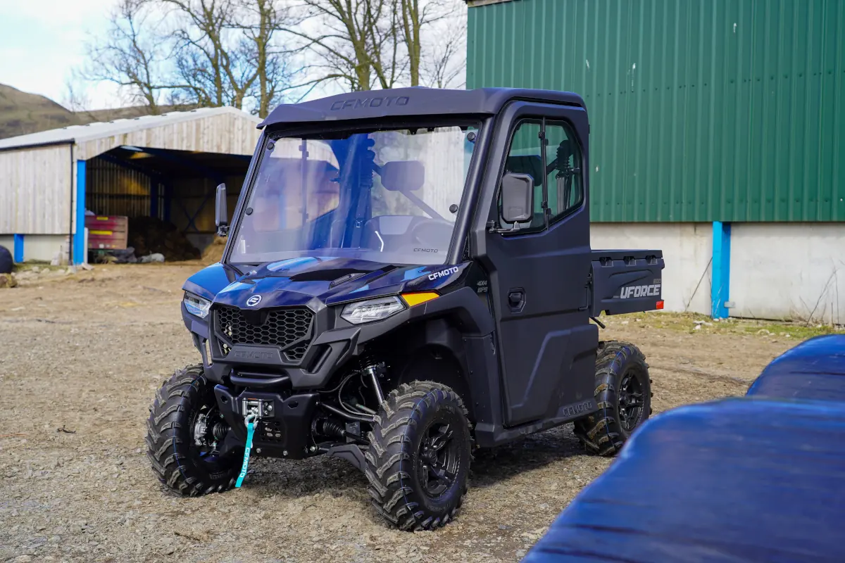 A black CFMOTO UFORCE 600 utility vehicle is parked on a gravel lot near green and blue farm buildings, with trees visible in the background.