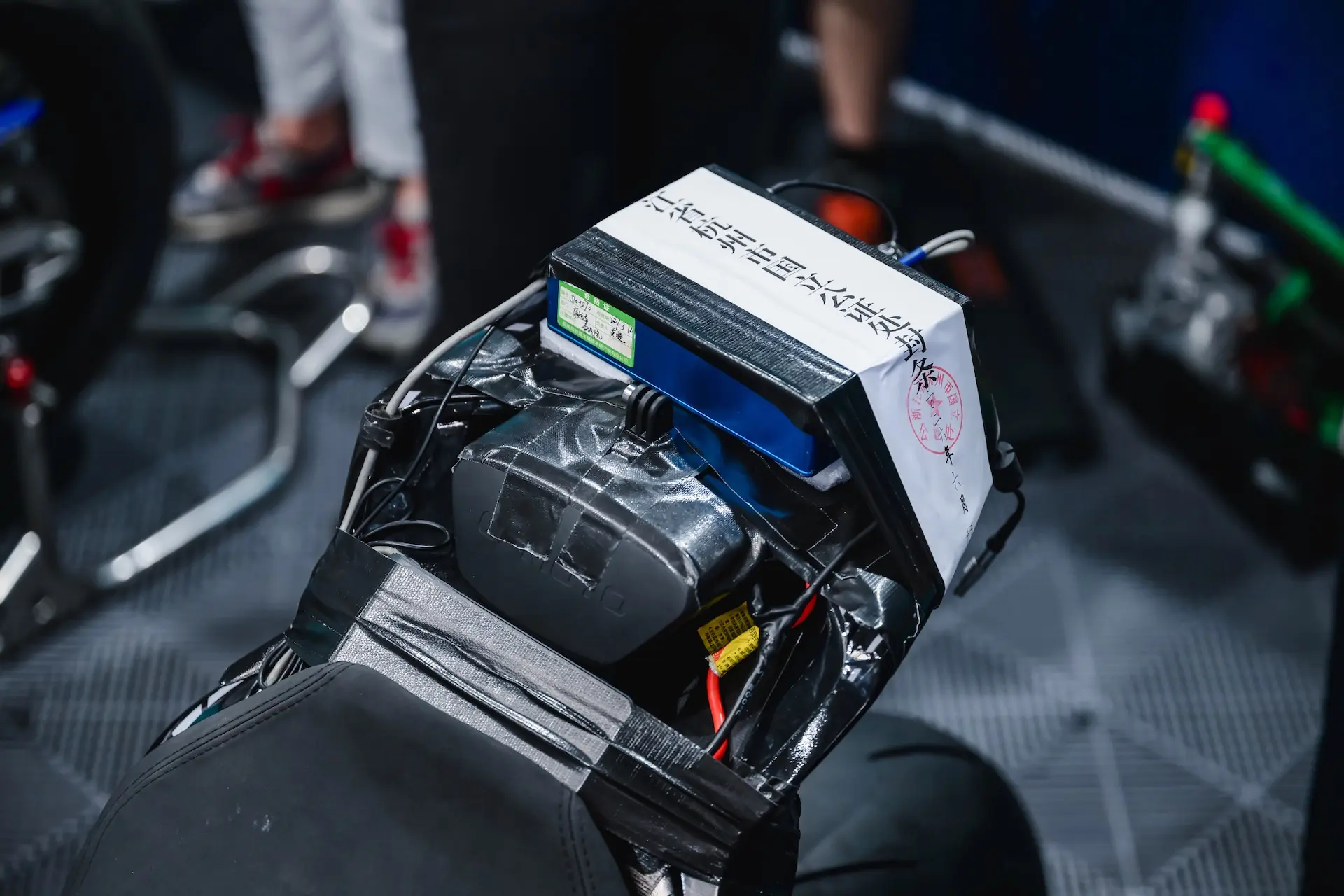 Close-up of a CFMOTO V4 SR-RR motorcycle seat with wiring, battery, and a white electronic device secured on top, surrounded by people in the background—highlighting its impressive 315.82 km/h top speed.