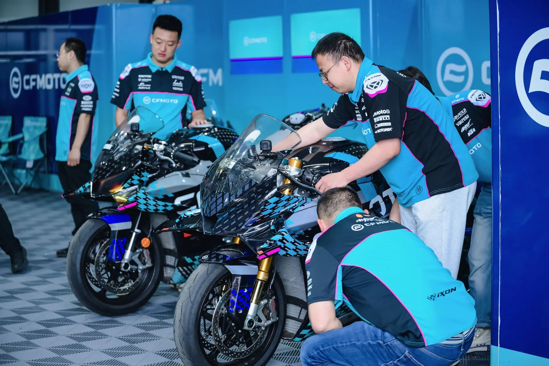 Four team members in matching uniforms prepare sport motorbikes, including the CFMOTO V4 SR-RR, in a garage area with blue CF MOTO branding visible in the background, aiming to achieve top speed on the track.