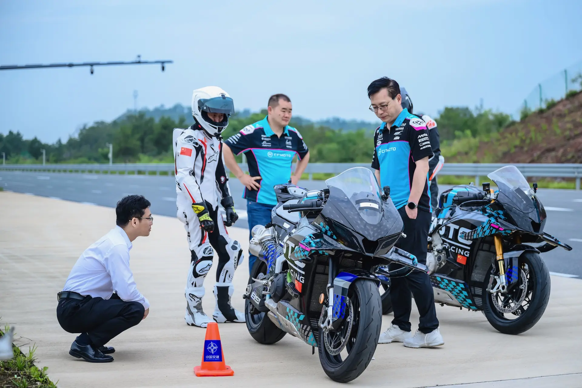 Four people, including a motorcycle racer in a white and red suit, stand with two CFMOTO V4 SR-RR racing motorbikes—machines capable of reaching a top speed of 315.82 km/h—on a paved surface outdoors.
