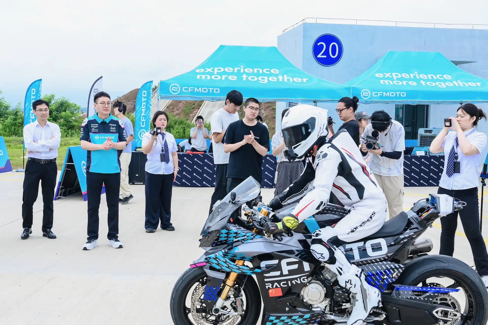 A motorcyclist in racing gear sits on a CFMOTO V4 SR-RR, known for its top speed of 315.82 km/h, while several people, some with cameras, observe and take photos at an outdoor event with branded marquees.