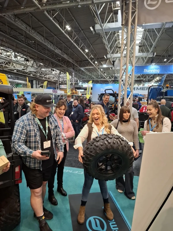 A woman lifts a large off-road tire with both hands at a busy trade show during its biggest and best year yet, surrounded by onlookers and various equipment displays.
