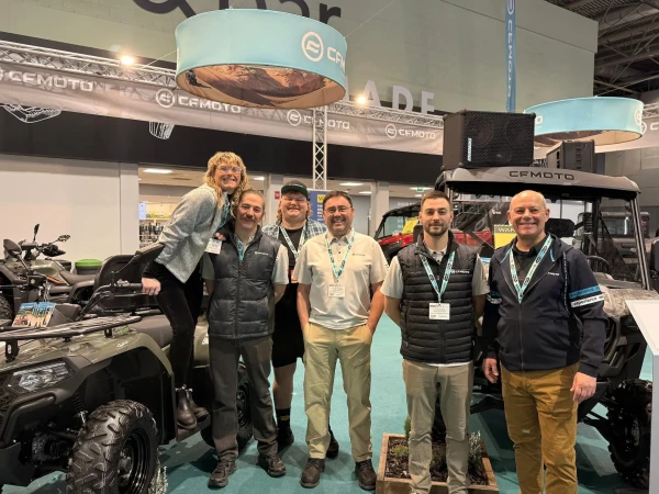 Six people stand and smile together at a CFMOTO exhibition booth, celebrating what promises to be their biggest and best year yet, surrounded by off-road vehicles and vibrant display banners.
