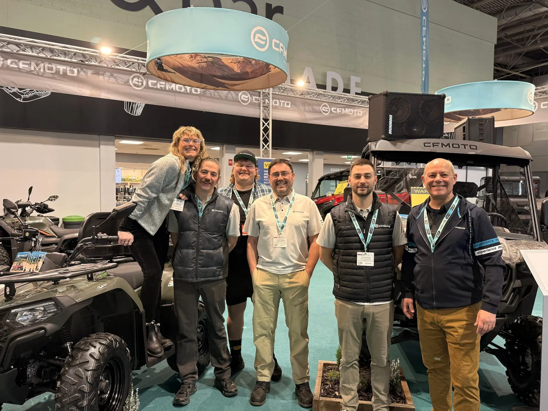 Six people stand and smile together at a CFMOTO exhibition booth, celebrating what promises to be their biggest and best year yet, surrounded by off-road vehicles and vibrant display banners.